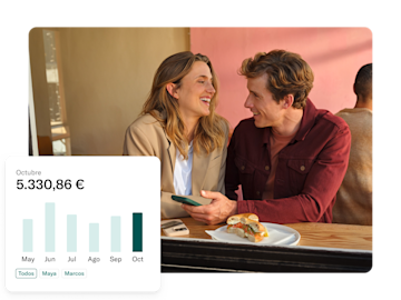Una pareja sonriente está sentada en una mesa con un sándwich, y una superposición de pantalla de teléfono muestra un gráfico de gastos mensuales de octubre con un saldo de 5.330,86€.