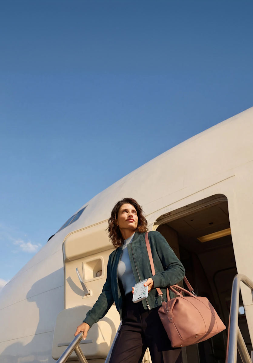 Una mujer con un bolso de viaje rosa baja por la escalerilla de embarque de un avión blanco mientras sostiene un teléfono inteligente.