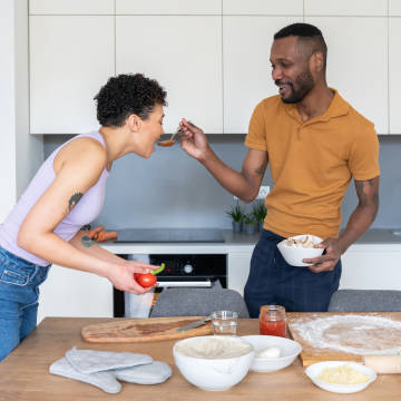 Imagen que muestra a un hombre y una mujer cocinando pizza juntos, mujer probando la salsa.