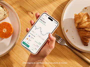 Una mano sostiene un smartphone que muestra un portafolio de criptomonedas sobre una mesa de madera con comida de desayuno.