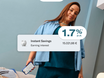 Woman dressed in blue, sitting, and holding her cell phone with the N26 application open. In the foreground, image of the 1.7% p.a. interest rate, balance in the main account, and in the savings account.