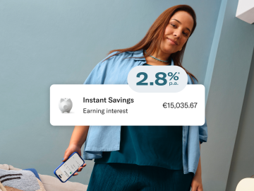 Woman dressed in blue, sitting, and holding her cell phone with the N26 application open. In the foreground, image of the 2.8% p.a. interest rate, balance in the main account, and in the savings account.