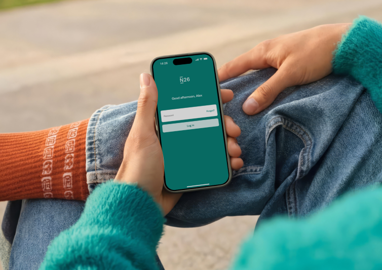 A person wearing a bright teal fuzzy sweater and blue jeans holds a smartphone displaying the N26 app login screen while sitting outdoors.