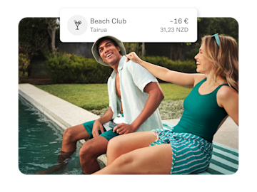 Un hombre y una mujer están sentados junto a una piscina, y una burbuja de notificación sobre ellos muestra una transacción para "Beach Club Tairua" por -16€ y 31,23 NZD.