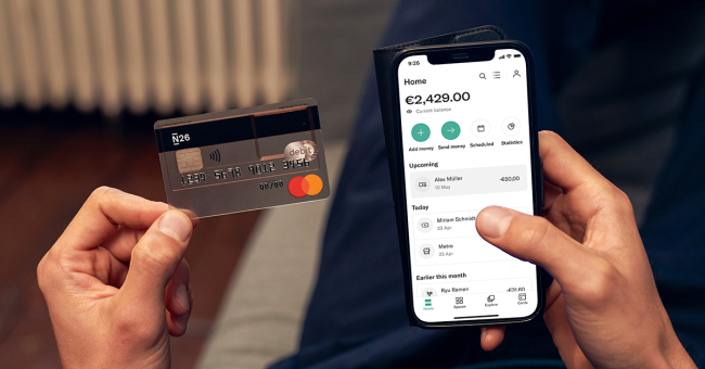 A hand holding an N26 standard card with a smartphone showing the N26 app.