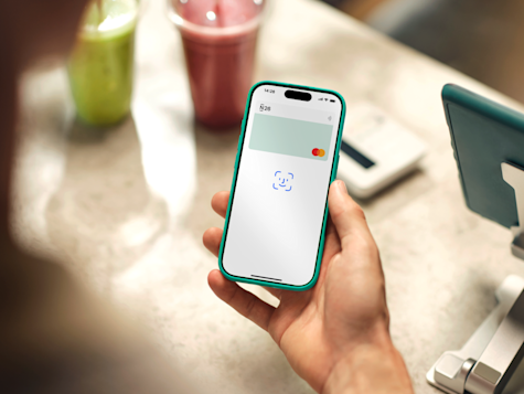 Eine Person hält ein Smartphone in einer türkisblauen Hülle über einen Tresen und zeigt die digitale Wallet-App von N26 mit einer virtuellen Mastercard, die neben zwei Smoothies zur Zahlung bereit ist.