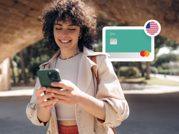 Imagen de una mujer joven parada en un parque y mirando su teléfono, con una tarjeta n26 y una bandera de Estados Unidos al fondo.