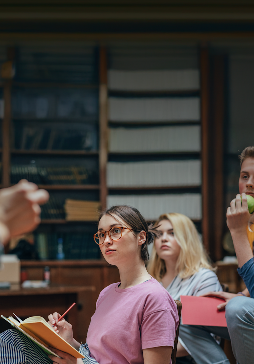 Eine junge Studentin mit Brille macht sich Notizen und hört aufmerksam während einer Vorlesung in einem schwach beleuchteten Klassenzimmer zu.