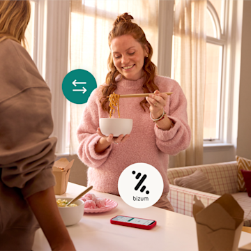 Una mujer sonríe mientras come fideos para llevar con una amiga en casa, con un smartphone en la encimera y los logotipos de transferencia de dinero y Bizum superpuestos.