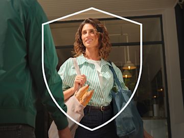 A smiling woman in a green striped shirt carries reusable grocery bags filled with fresh produce and baguettes while walking out of a store, with a shield-shaped outline symbolizing security in the foreground.