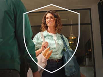 Una mujer sonriente con una camisa de rayas verdes lleva bolsas reutilizables llenas de productos frescos y baguettes mientras sale de una tienda, con un contorno en forma de escudo en primer plano que simboliza la seguridad.