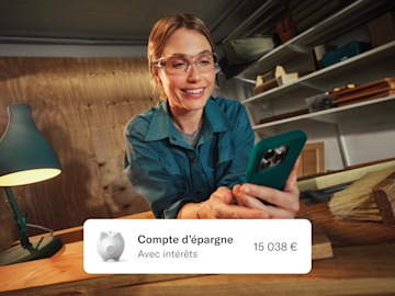 Une femme souriante portant des lunettes de protection regarde son smartphone dans un atelier, avec une carte superposée affichant un solde d'épargne.
