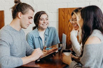 Three women and a man having a stand up conversation and drinking coffee.