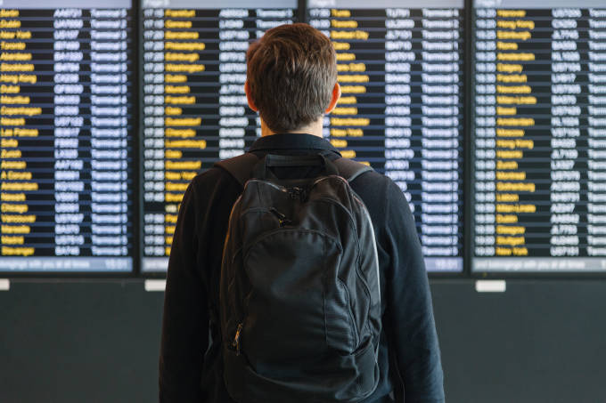 Junger Mann mit Rucksack vor Abflugtafel am Flughafen.