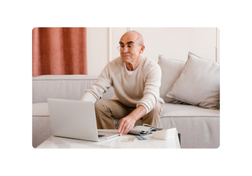 Un uomo anziano con gli occhiali è seduto su un divano e usa un computer portatile. Sul tavolo davanti a lui c'è un misuratore di pressione sanguigna e delle pillole.