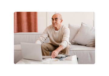 A man with glasses sits on a couch, his laptop open. On the table before him, a blood pressure cuff and medication await his attention.