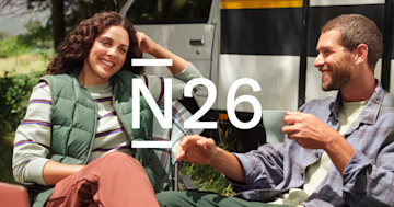 A smiling man and woman sit outdoors near a camper van while enjoying drinks, with the N26 logo overlaid in the center.
