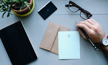 Man holding a pencil on a table with glasses, notebook, and envelopes.