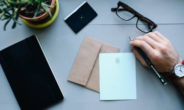 Hombre que sostiene un lápiz sobre una mesa con gafas, cuaderno y sobres.