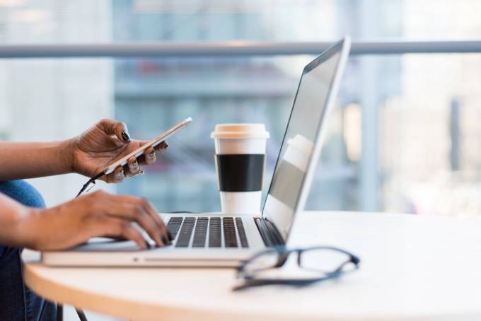 Person working on a table with a laptop, smartphone, pair of glasses and a cup of coffee on it.