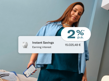 Woman dressed in blue, sitting, and holding her cell phone with the N26 application open. In the foreground, image of the 2% p.a. interest rate, balance in the main account, and in the savings account.