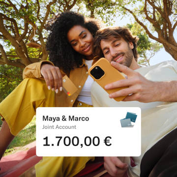 Maya and Marco sitting in a Park. Maya is holding her N26 card and Marco looking at his mobile. In the foreground there is a pop up with the amount of money in their joint account.
