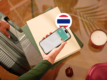 image of a person holding a phone showing an N26 card in the app screen and grabbing a suitcase with the other hand and with a flag from Thailand.