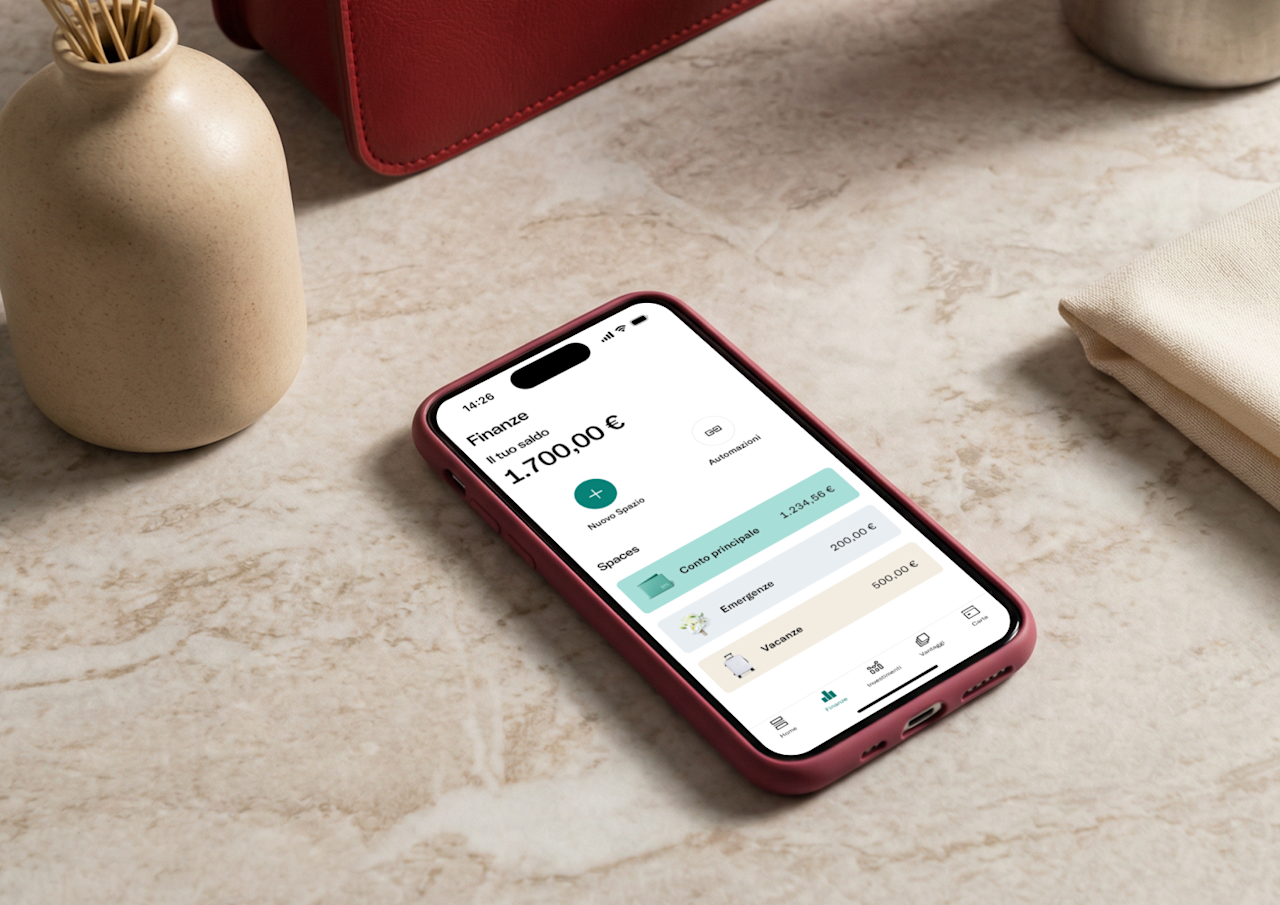 Uno smartphone rosa su una superficie di marmo mostra la schermata delle finanze dell'app N26 con un saldo di 1.700,00 € e sottoconti categorizzati per fondi di emergenza e vacanze.