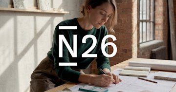  A woman drafts architectural plans at a wooden table in a brightly lit studio with a large white N26 logo overlaid in the foreground.