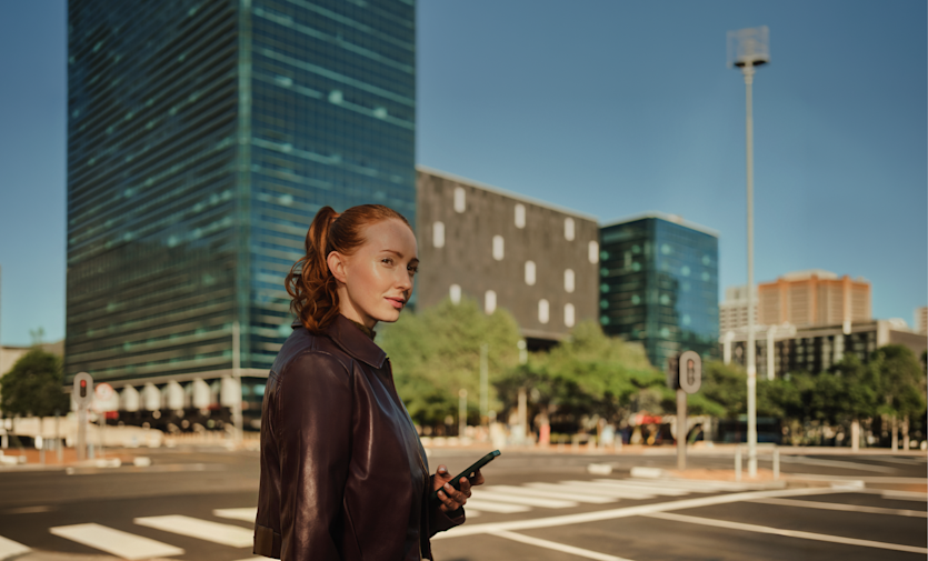 Eine Frau mit zu einem Pferdeschwanz gebundenen roten Haaren hält ein Smartphone, während sie an einem Fußgängerüberweg in einer Stadt mit modernen Bürogebäuden steht.