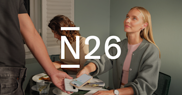 Una mujer realiza un pago sin contacto con su teléfono a una persona que sostiene un datáfono en una mesa de restaurante, con el logotipo de N26 superpuesto.