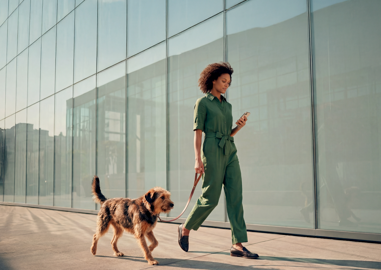 Une femme aux cheveux bouclés portant une combinaison verte regarde son smartphone tout en promenant son chien le long d'un bâtiment moderne en verre.