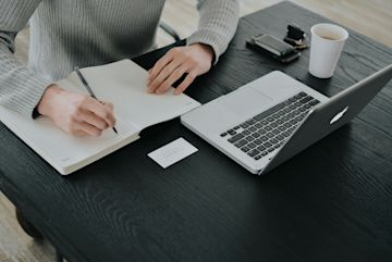A person writing in a notebook, looking at a laptop.