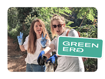 Two women wearing gloves and holding trash bags smile and pose for the camera in a wooded area, with the words "GREEN ERG" overlaid on the image.