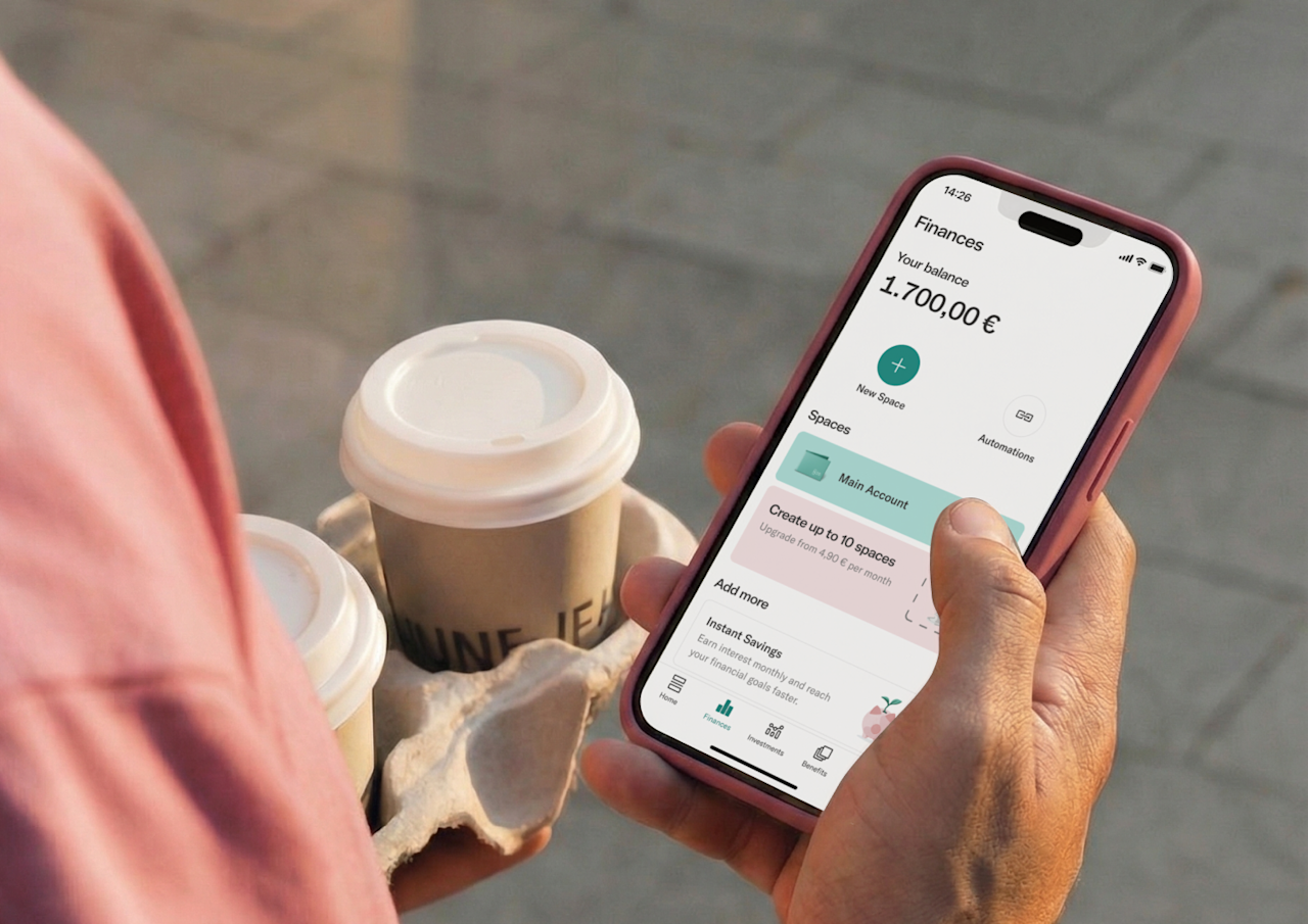 A close-up, over-the-shoulder view of a person holding a tray of takeaway coffees in one hand while checking a mobile banking app on their smartphone with the other.
