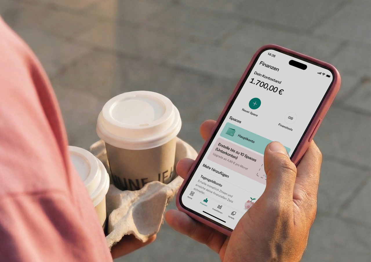 Eine Nahaufnahme über die Schulter einer Person, die in einer Hand ein Tablett mit Kaffees zum Mitnehmen hält, während sie mit der anderen eine mobile Banking-App auf ihrem Smartphone prüft.