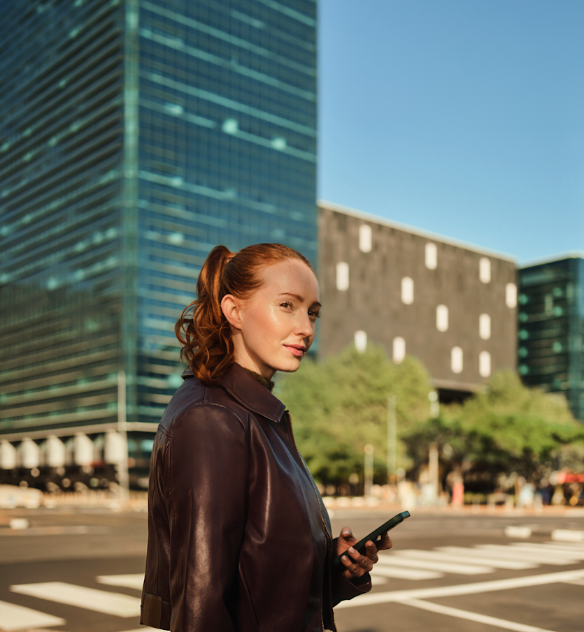 Eine Frau mit zu einem Pferdeschwanz gebundenen roten Haaren hält ein Smartphone, während sie an einem Fußgängerüberweg in einer Stadt mit modernen Bürogebäuden steht.