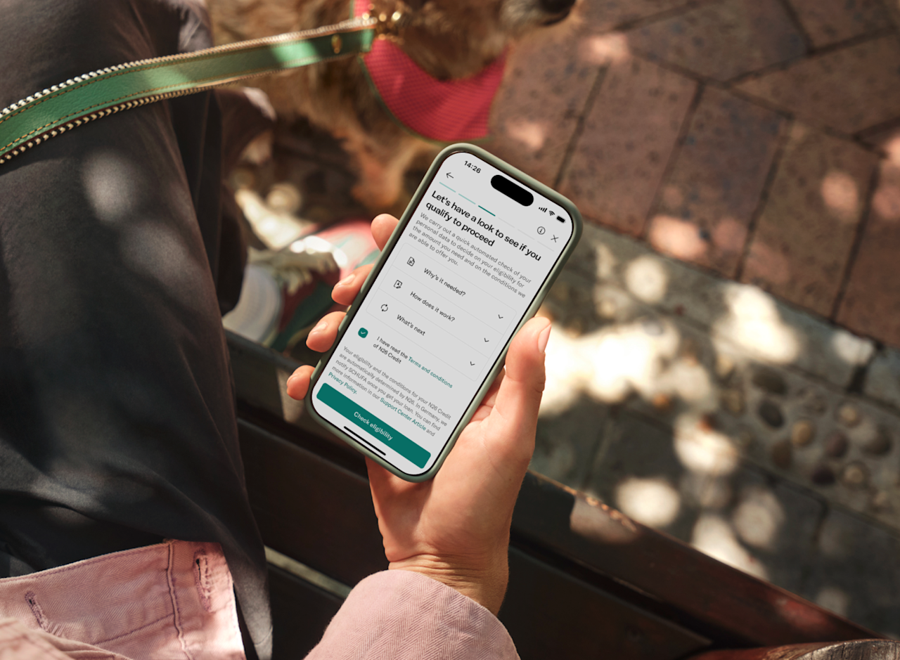 A hand holds a smartphone displaying the N26 screen for checking loan eligibility, with a dog visible in the background.