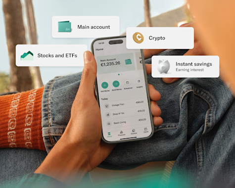 A person holds a smartphone showing a financial app with features like "Main account," "Stocks and ETFs," "Crypto," and "Instant savings" highlighted. The background includes a casual outdoor setting with jeans and bright orange socks.
