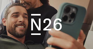 A couple sits smiling on the sofa, the man looks at his mobile phone.