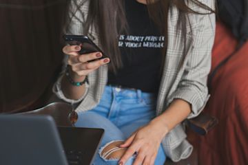 A woman by a laptop looking at her cellphone.