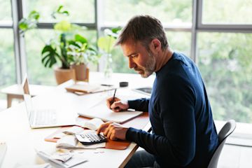 Man taking notes working with a calculator and a laptop.