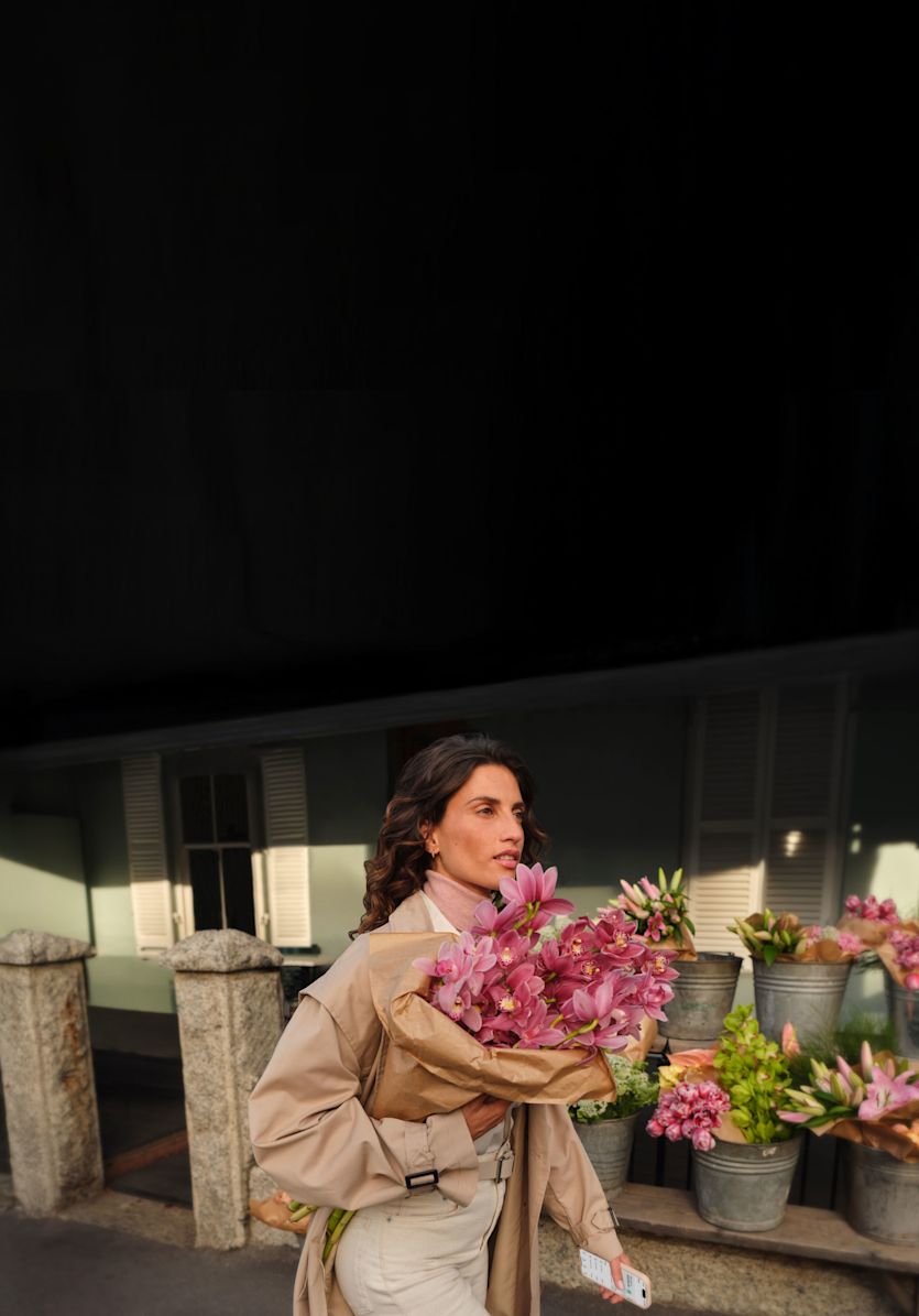 Una mujer con un abrigo beige sostiene un ramo de flores rosas y un teléfono inteligente mientras camina junto a una floristería.