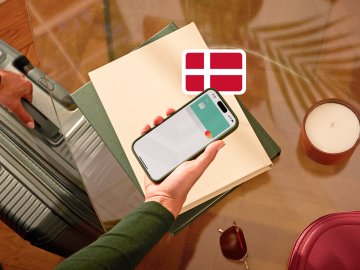 image of a person holding a phone showing an N26 card in the app screen and grabbing a suitcase with the other hand and with a Danish flag.