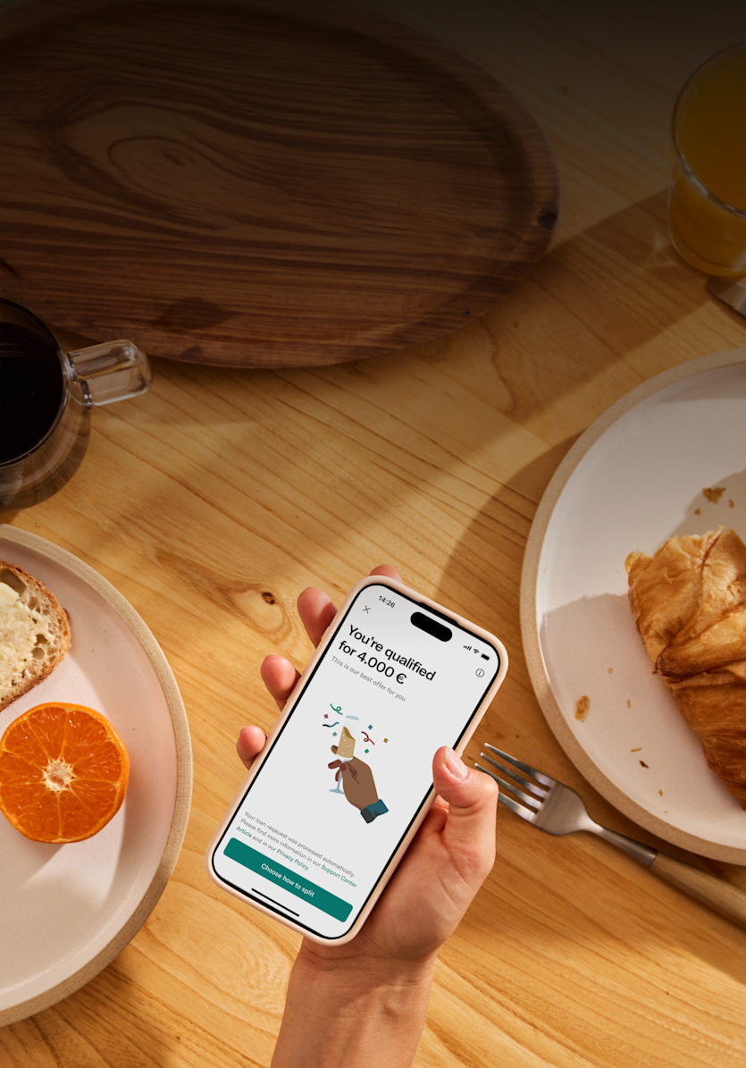 An overhead shot of a hand holding a smartphone displaying a loan offer on a wooden table with breakfast food.