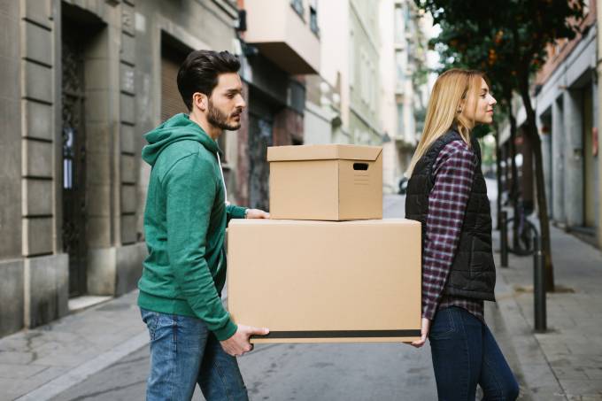 a young woman and a young man crossing the street carrying moving boxes.