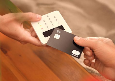 Ein Nahaufnahmefoto zeigt eine Person, die eine schwarze N26 Mastercard hält, während eine andere Person ein kleines, weißes Kartenlesegerät mit Tastenfeld und Display in der Hand hat. Die Karte wird gerade in das Lesegerät eingeführt oder in dessen Nähe gehalten, um eine Transaktion durchzuführen. Der Hintergrund ist weich verschwommen und hebt die Hände, die Karte und das Lesegerät hervor. Die Szene vermittelt den Eindruck einer einfachen und modernen Bezahlmethode.