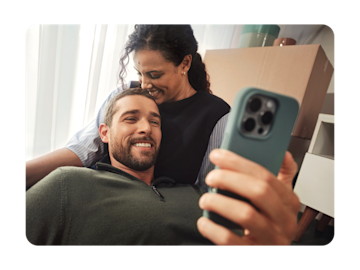A couple is sitting on their sofa, he is looking at his phone and both are smiling.