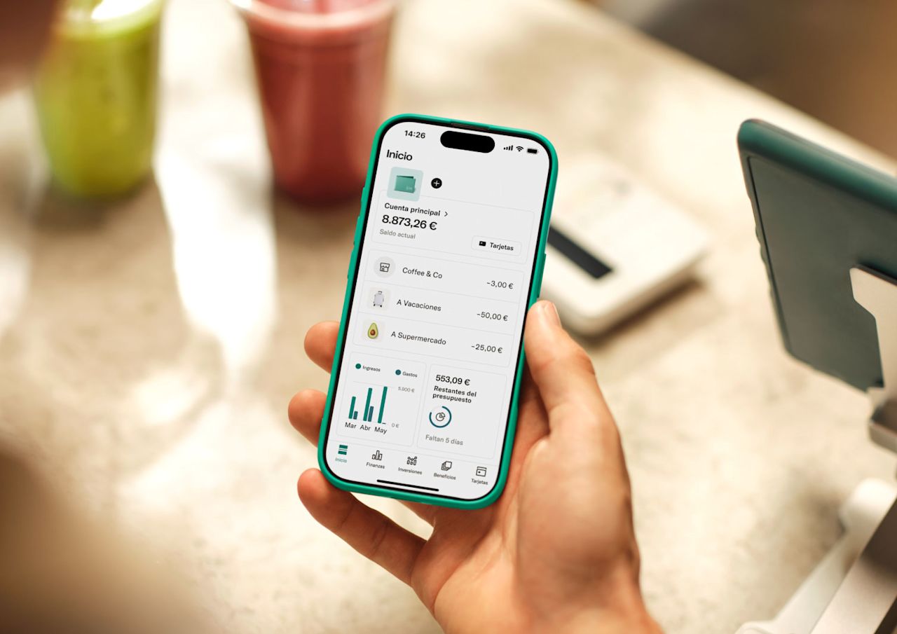 Una persona sostiene un smartphone que muestra la aplicación bancaria N26. La aplicación muestra el saldo de la cuenta principal de **8.873,26 €** y transacciones recientes como "Coffee & Co." y "To Travel.".