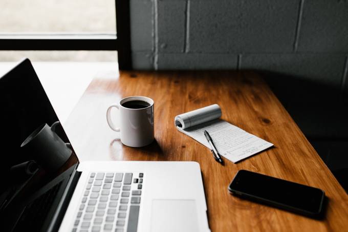 Un bureau en bois avec un ordinateur portable, un carnet, une tasse et un smartphone.